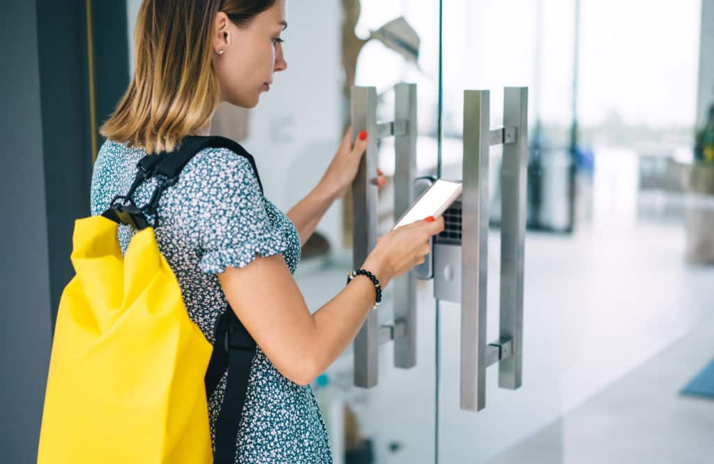 Pensive blonde woman in casual wear with backpack open door using smartphone with mock up screen application, serious female unlocking security system via mobile phone checking code for entrance AdobeStock 336979669